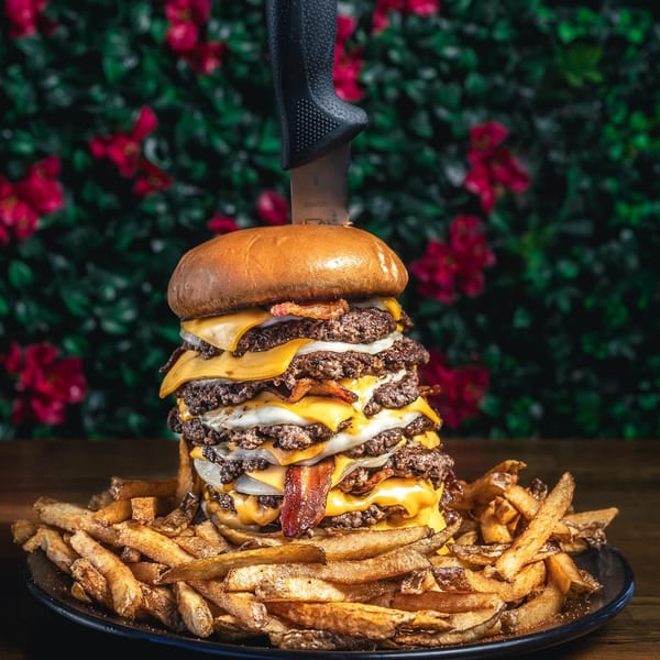 A towering Chelito's burger with multiple patties, cheese, bacon, and onions, served with fries.
