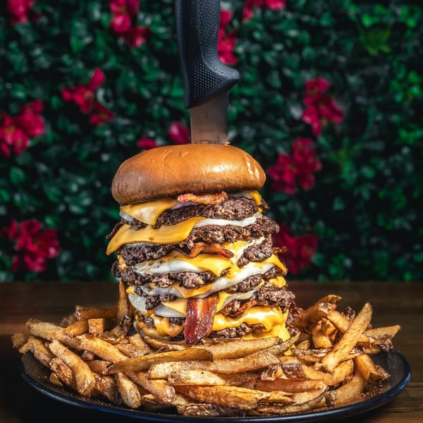 A massive Chelito's burger with multiple patties, cheese, bacon, and onions, served with fries.