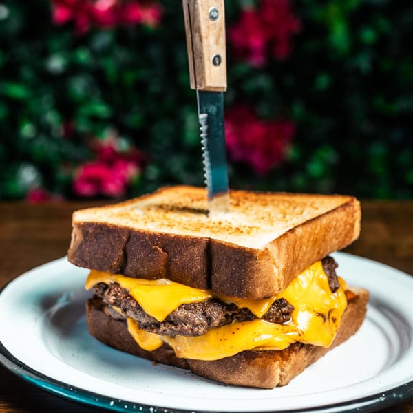 Chelito's double cheeseburger with melted cheese between toasted bread, held by a knife, on a white plate.