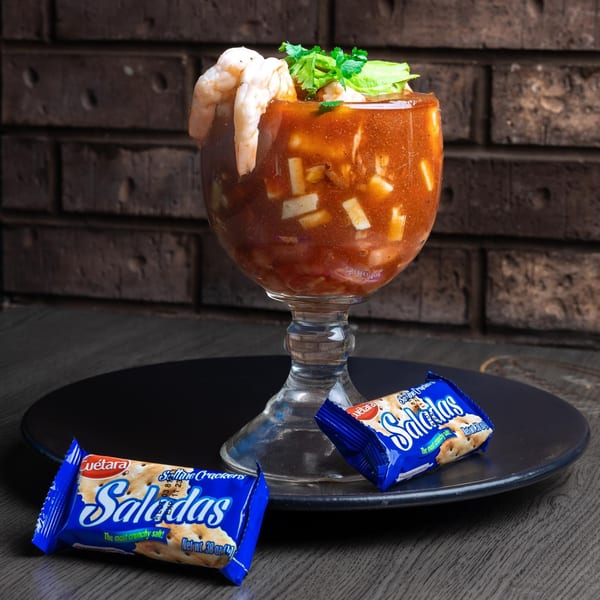 A shrimp cocktail in a goblet, garnished with avocado and cilantro, with two cracker packets.